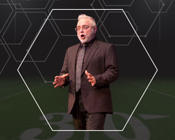 Person wearing a dark suit and tie standing on a football field background with hexagonal graphic overlay.